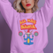 A Christmas sweatshirt featuring a playful design with a cartoon Baphomet goat character wearing a Santa hat. The design includes the text "All Hail Santa" above the goat, with pentagrams in flames on either side. The goat is depicted in a meditative pose, adding a humorous and irreverent twist to holiday attire.